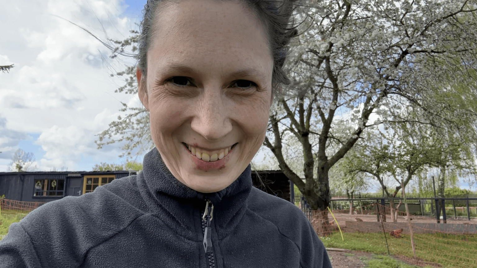 A portrait of Kate, grinning. She has some wounds from cold sores on her lips. Behind her is a blossomig cherry tree.