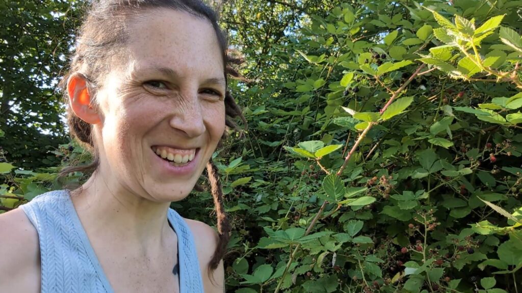 Kate is smiling next to blackberry brambles with fruit.