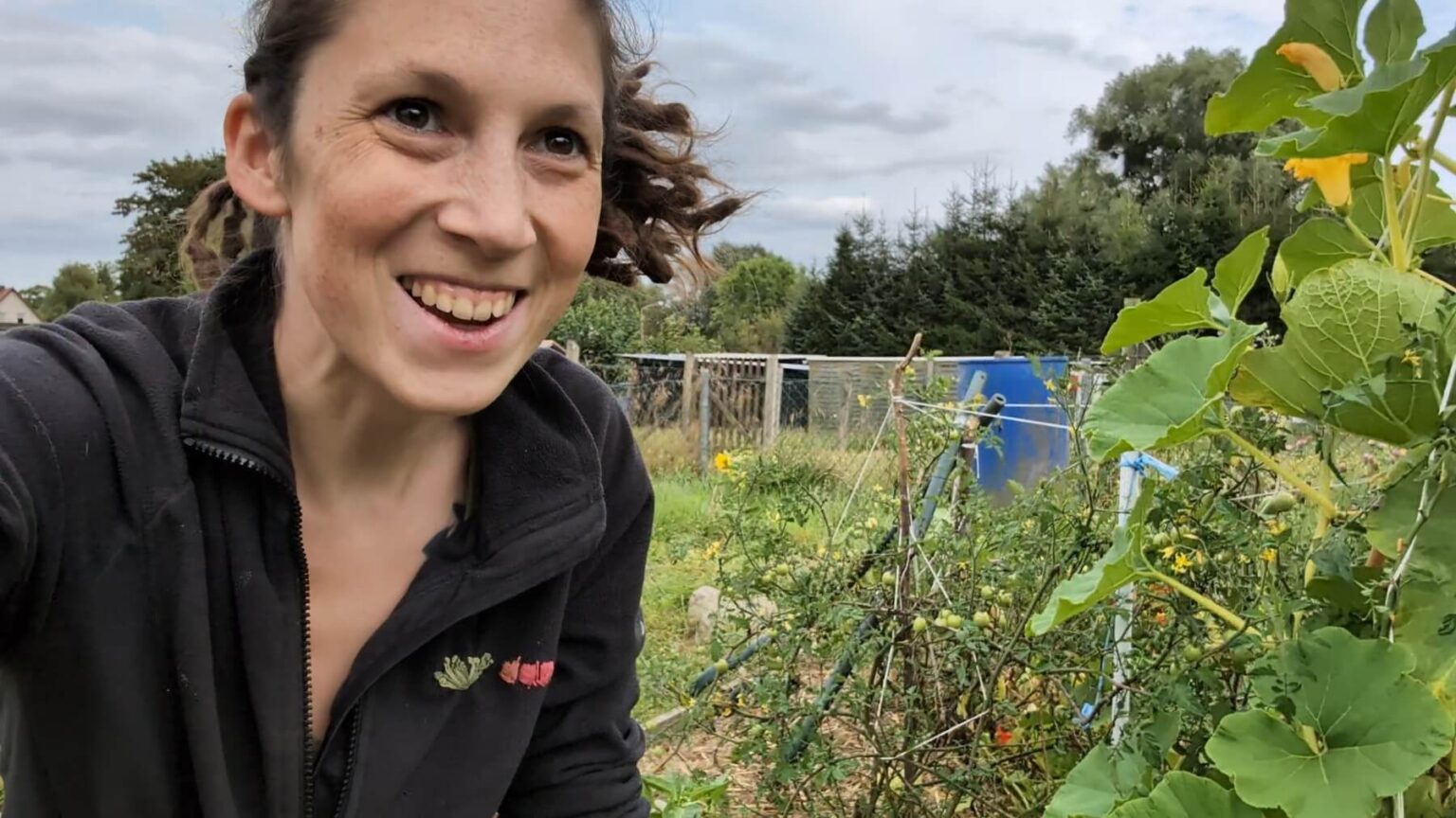 Kate is smiling at something off-camera. Next to her a pumkin grows up a trellis. In the background are tomatoes.