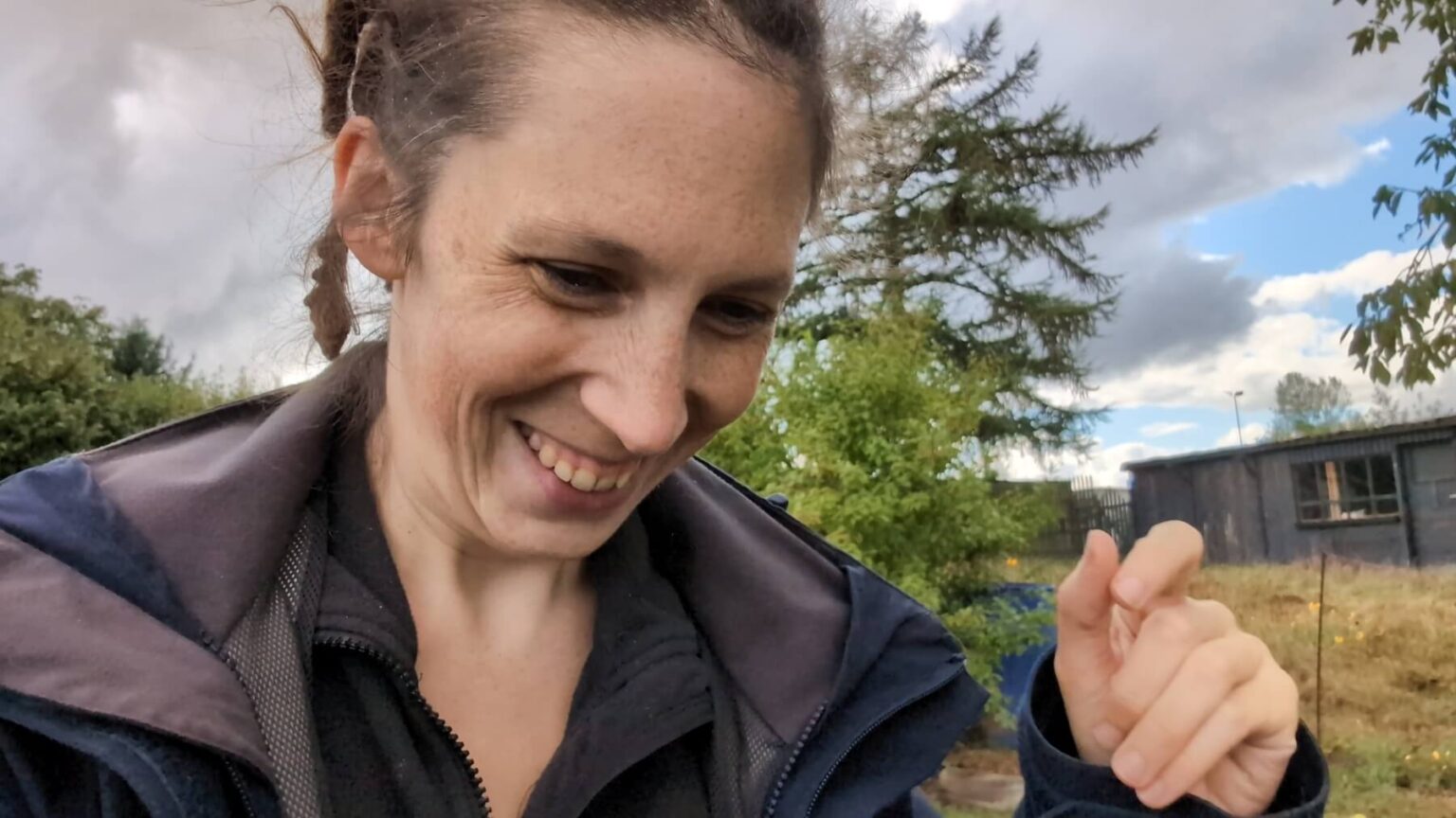 Kate is grinning widely in front of a stormy sky. She is wearing a rain jacket.