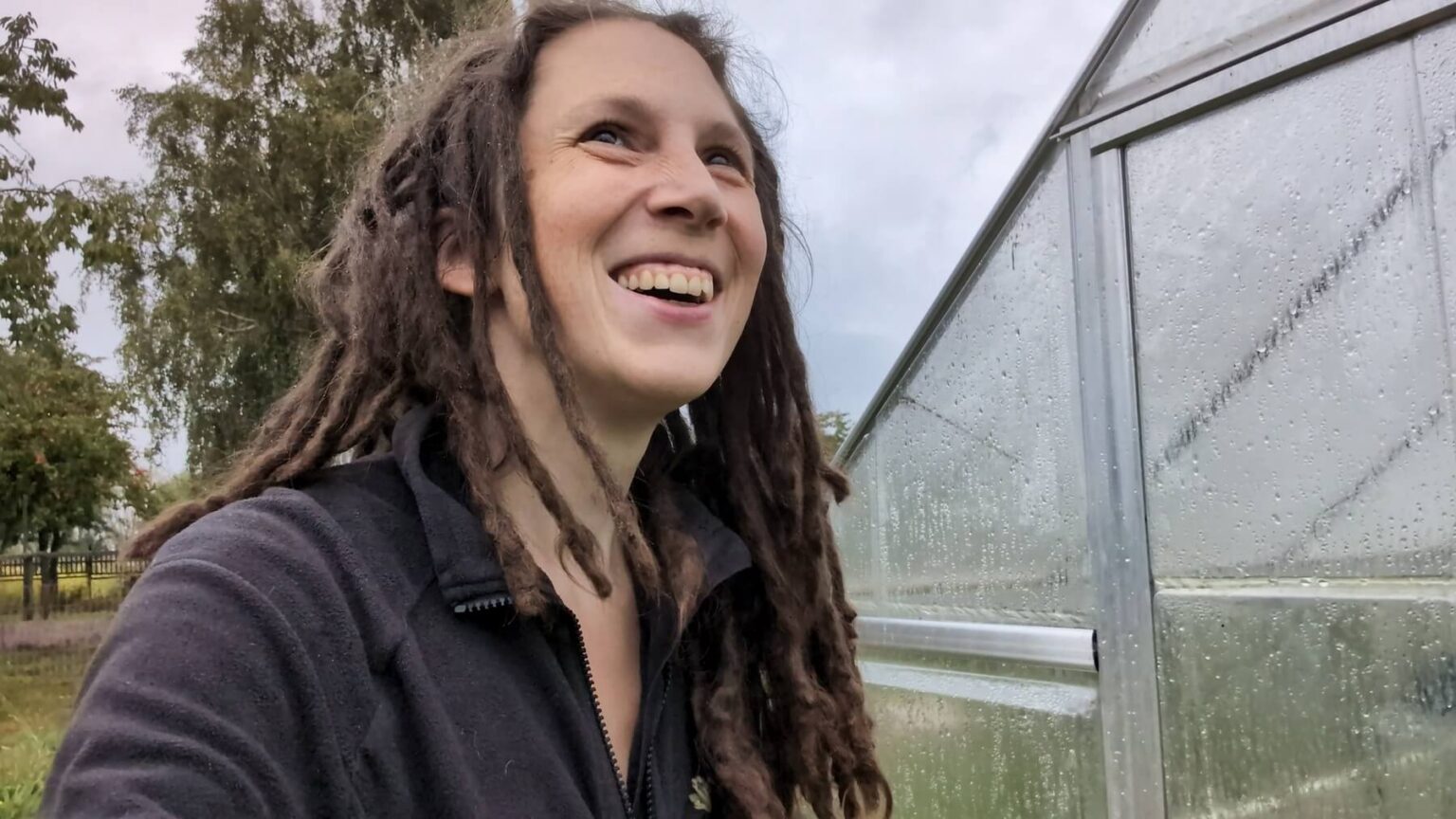 Kate is grinning up at the sky despite more rain. The greenhouse next to her is covered in drops.