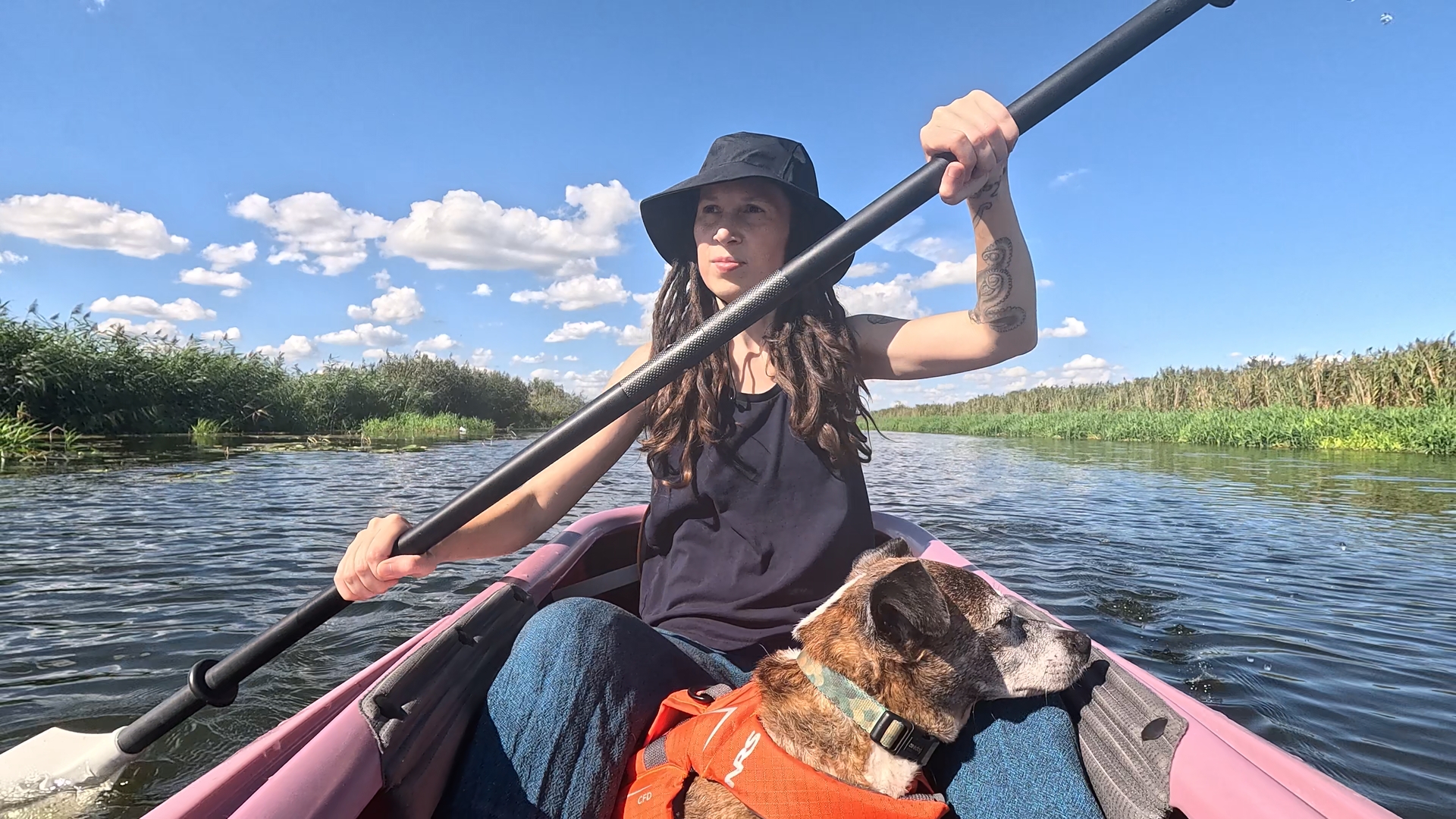 Kate in einem Kajak auf einem Fluss mit Schilf-Ufer. Pepper liegt vor ihrem Schoßmit seinem Kopf auf Kates Knie.
